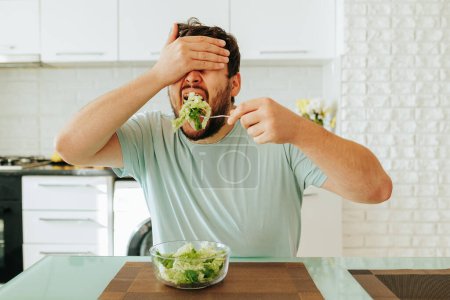 Front view, closing his eyes with his hand, the young man brought a fork with salad to his mouth. He reluctantly eats healthy food. Concept of diet food. There is a glass bowl on the table. Stop diet.