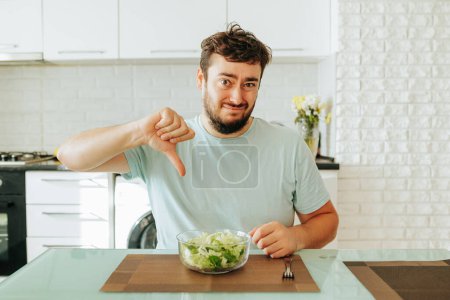 A young man sits in the kitchen in front of a glass bowl of salad and shows a dislike with a twisted face. The guy doesnt want to eat healthy food anymore. Modern interior. Stop diet.