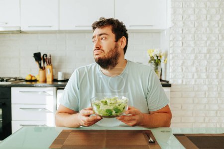A young man guy holds a bowl of salad in his hands and looks doomed into the distance. A man sits at a table in his kitchen and does not want to eat healthy food. Curves. Stop diet.
