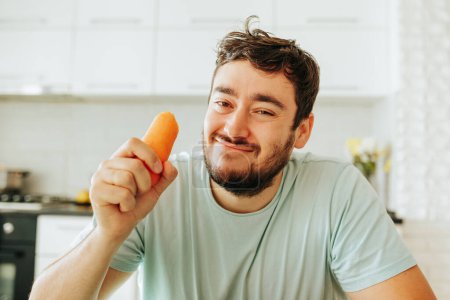 Smiling derisively at the camera, a young man holds a raw orange carrot in his hand. Maybe he is on a diet to improve his health. The guy does not want to eat it.