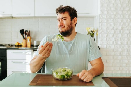 Frustrated by the need to eat unloved food, a young guy holds a fork with a green salad in his hand. Diet food is not what the guy dreamed of. Stop diet, healthy food, vegan food.