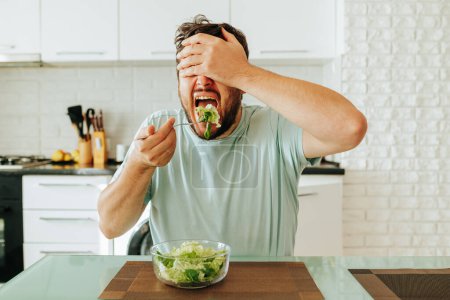 With mouth wide open young guy man brings fork with salad to his mouth, closing his eyes with his hand. Healthy diet, raw vegetables and greens. It is useful to monitor health and weight. Stop diet.