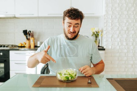 Sentado en la cocina mirando en un tazón de ensalada un hombre piensa lo cansado que está de estilo de vida saludable. El joven quiere estar sano, pero es duro y no divertido. Curvas caras, mostrando diferentes emociones.
