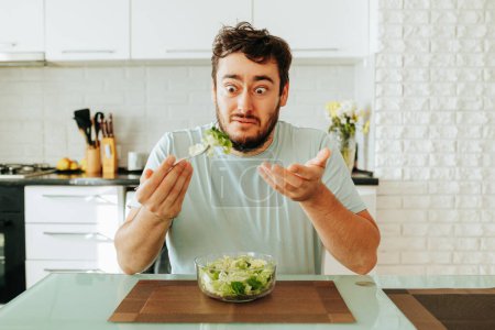 Front view young guy man looks at a fork with salad and hates vegetables. Juicy bright ripe salad lies in a bowl. Modern interior. Healthy food. Healthy lifestyle. Stop diet.
