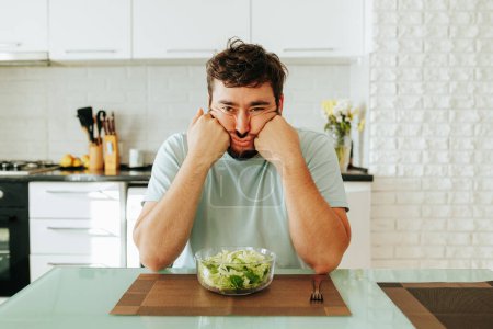 A glass bowl with a delicious juicy vitamin salad stands in front of an unhappy young man who is forced to diet. The young man holds his head in his hands and looks hopelessly ahead. Stop diet.