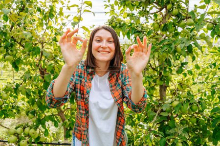 Sehr glückliche junge Bäuerin Agronomie gestikulierend, dass alles cool ist. Blick von vorne auf die Kamera Obstbäume Obstgarten. Viel Grün. Das Konzept von Landwirtschaft und Glück.