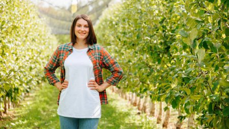 Looking at camera stand among rows of fruit trees young farmer woman hands hips smiling joyful. Agronomy adult girl is glad harvest rich. Family garden, family business, young business is successful.
