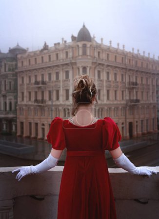 Schöne junge Mädchen in einem roten Kleid aus dem neunzehnten Jahrhundert steht auf einem Balkon vor einem schönen Gebäude.