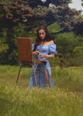 Foto de Hermosa joven mujer es relajante mientras que la pintura de un lienzo de arte al aire libre en el parque bajo la lluvia. - Imagen libre de derechos