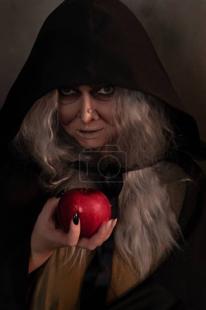 Foto de La madrastra bruja da manzana roja envenenada.La mujer como bruja en negro ofrece manzana roja como símbolo de tentación, veneno. Cuento de hadas, concepto de mago de nieve blanca. Halloween espeluznante, cosplay. - Imagen libre de derechos