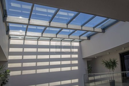 Téléchargez les photos : Vue en perspective du hall moderne verre et plafond en acier avec de forts motifs géométriques et des ombres sur le mur comme fond abstrait avec de l'espace pour la copie. Plantes vertes, ciel bleu - en image libre de droit