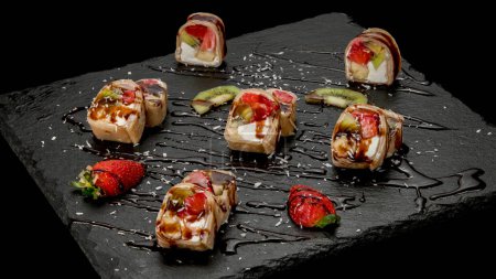 Téléchargez les photos : Rouleaux de crêpes au chocolat maki sushi farcis de fruits et fromage close-up. Crêpe de dessert tranchée à la crème fouettée, kiwi et fraise sur une élégante assiette de restaurant en pierre sur fond noir - en image libre de droit
