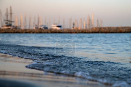 Téléchargez les photos : Belle vue à angle bas des vagues bleues qui roulent sur le rivage sablonneux au coucher du soleil. Mâts de yachts amarrés dans le port sur un fond flou, faible profondeur de champ - en image libre de droit