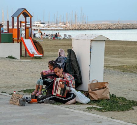 Téléchargez les photos : Larnaca, Chypre - avril 01, 2024 : Triste femme sans abri avec une petite fille, musicien de rue jouant de l'accordéon sur le trottoir avec une plage de sable sur fond flou. Petit seau d'aumônes bosselées, signe "affamé" - en image libre de droit