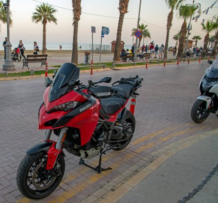Téléchargez les photos : Larnaca, Chypre - 01 avril 2024 : Moto Red Ducati Multistrada garée dans une rue avec des palmiers et de la mer en arrière-plan - en image libre de droit