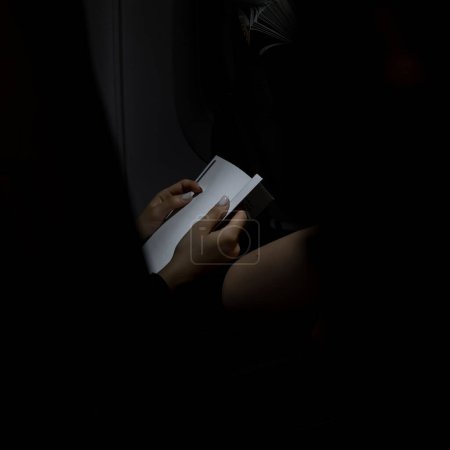 Téléchargez les photos : Jeune femme méconnaissable lisant le livre dans l'avion pendant le vol de nuit. Scène contrastée où seuls le livre et une partie du corps des filles sont illuminés dans le noir. Concept vacances et voyages - en image libre de droit