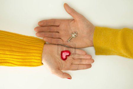 Two hands, male and female, extend their palms to each other. In one hand is a red heart, in the other hand is a small key. Close-up. Isolated on white background.