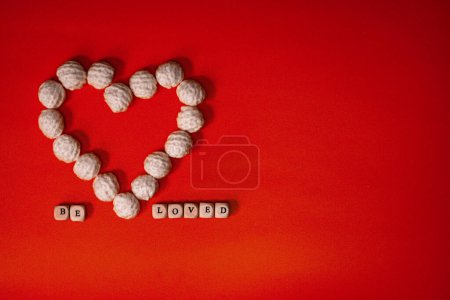 Foto de A white heart made of knitted balls and the inscription be loved from white wooden cubes on a red vintage background. Place for text. - Imagen libre de derechos