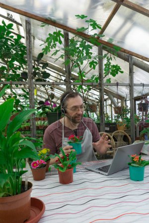 Photo for Man in an apron sells flowers and seedlings via video link on a laptop via the Internet. He has a headset on his head for conversations. He holds flowers in his hands and smiles. Gardening, sale - Royalty Free Image