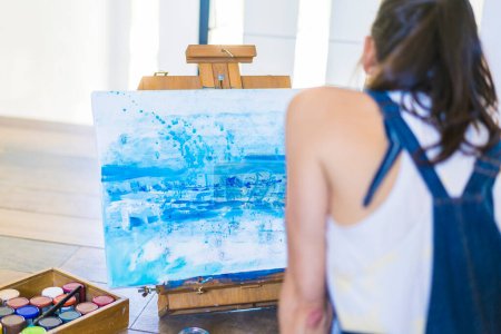 Eine Künstlerin malt ein blaues abstraktes Gemälde auf eine Leinwand. Kreative junge Frau arbeitet auf dem Fußboden in ihrem Kunstatelier.