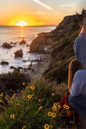 Téléchargez les photos : Vue arrière de jeunes femmes regardant le coucher du soleil dans les collines d'El Matador Beach, Malibu, Californie - en image libre de droit