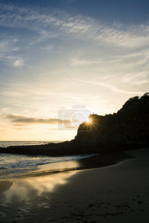 A beautiful sunset over the ocean with a bright sun shining on the water on a rocky beach