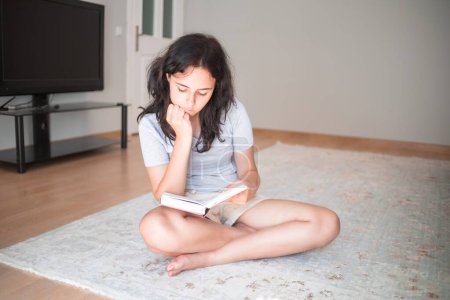 Téléchargez les photos : Mignon jeune fille lecture livre de papier tout en étant assis sur le tapis à la maison - en image libre de droit