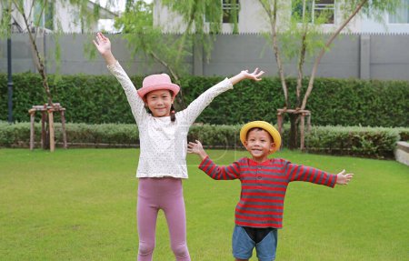Téléchargez les photos : Heureux petit garçon asiatique et jeune fille enfant portant un chapeau de paille debout et saluant au revoir avec caméra dans le jardin. - en image libre de droit