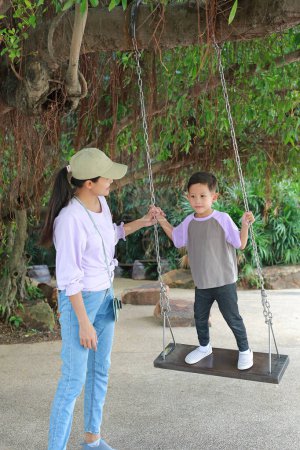 Téléchargez les photos : Bonne famille asiatique mère balançoire tandis que le petit garçon debout sur elle dans le jardin. Maman et fils jouent swing. - en image libre de droit