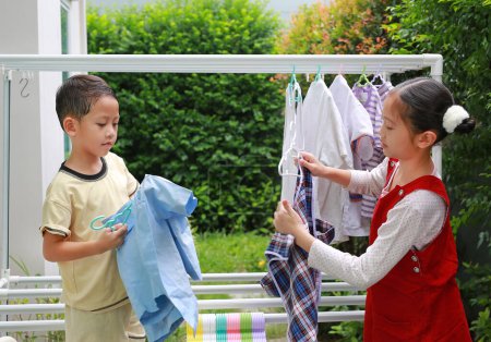 Téléchargez les photos : Asiatique fille enfant et petit garçon aider à faire sécher les vêtements dans le jardin près de la maison. Soeur et petit frère aident au ménage. Les enfants accrochent la blanchisserie sur un rail de corde à linge. - en image libre de droit