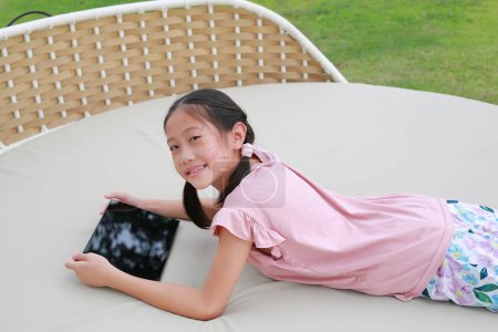 Téléchargez les photos : Souriant asiatique fille enfant jouer tablette et regarder la caméra tout en étant couché sur canapé en tissu dans le jardin. - en image libre de droit
