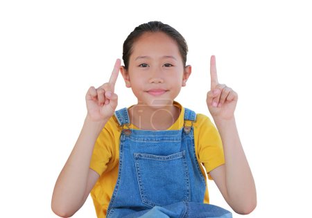 Téléchargez les photos : Gros plan de fille asiatique enfant en dungarees jean montrant deux index et caméra de regard isolé sur fond blanc. - en image libre de droit