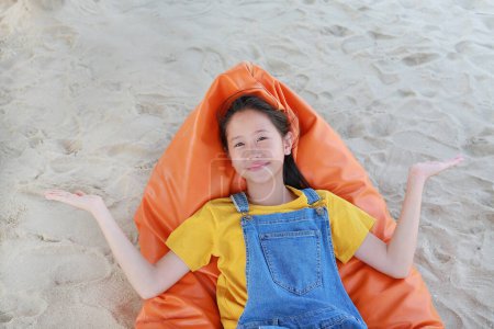 Retrato de niña asiática feliz en dungarees jean abrir las manos o extender los brazos y mirando a la cámara mientras se relaja en la playa sofá cama naranja en la arena en las vacaciones de verano.