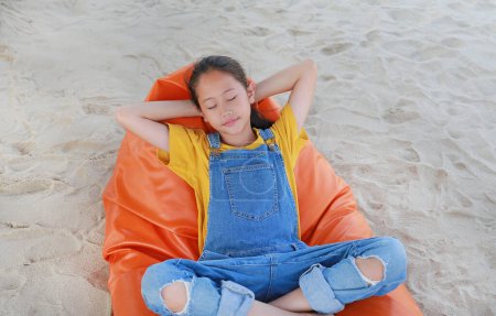 Téléchargez les photos : Souriant Asiatique fille enfant en dungarees jean se détendre et dormir sur le canapé-lit orange plage sur le sable pendant les vacances d'été. - en image libre de droit