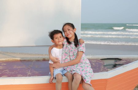 Téléchargez les photos : Portrait de heureux Asiatique garçon et fille assis et câlin tout en étant assis sur voilier et regardant caméra sur la plage pendant les vacances d'été. - en image libre de droit