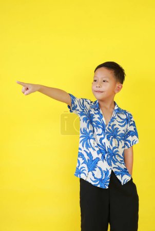 Téléchargez les photos : Portrait de garçon asiatique en costume d'été pointant index ou index et regardant à côté tout en se tenant debout sur fond de studio jaune. - en image libre de droit