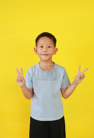 Téléchargez les photos : Asiatique petit garçon gosse montrant la victoire signe doigts et regarder droit tout en se tenant isolé sur fond jaune. - en image libre de droit