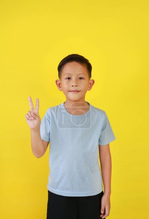 Téléchargez les photos : Portrait de garçon asiatique enfant montrent des doigts de signe de victoire et regardant droit tout en se tenant isolé sur fond jaune. - en image libre de droit