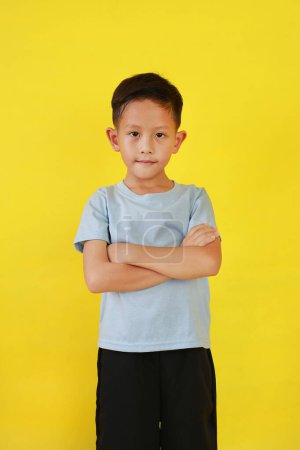 Téléchargez les photos : Portrait d'un garçon asiatique souriant âgé d'environ 7 ans debout et les bras croisés sur la poitrine regardant directement la caméra isolée sur fond de studio jaune. - en image libre de droit