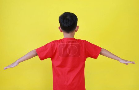Téléchargez les photos : Vue arrière de garçon asiatique les mains ouvertes les mains tendues. Enfant debout et écarter ses bras largement isolé sur fond jaune. - en image libre de droit