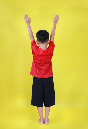 Téléchargez les photos : Vue arrière du petit garçon asiatique les mains levées. Enfant debout et levez les bras isolés sur fond jaune. Image pleine longueur. - en image libre de droit