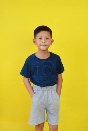 Téléchargez les photos : Souriant asiatique petit garçon enfant mettre la main dans sa poche de pantalon et regarder droit sur fond jaune studio. - en image libre de droit