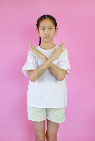 Porträt eines asiatischen Mädchens im Alter von 9 Jahren kreuzte seine Hände Geste isoliert auf rosa Studiohintergrund. Kind zeigt Stoppschild oder macht X-Zeichen Arme und schaut Kamera.