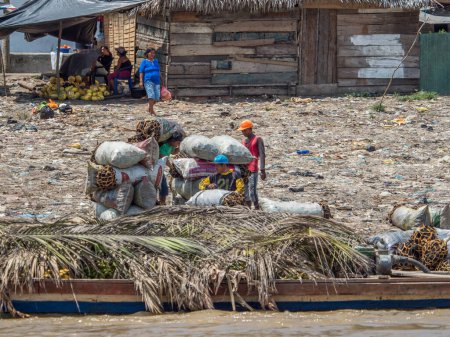 Téléchargez les photos : Iquitos, Pérou - 25 sept. 2018 : Les hommes portent les sacs avec du charbon de bois. Une énorme pollution peut être vue en arrière-plan sur la rive de la rivière Itaya. Basse saison des eaux de l'Amazonie. Quartier Beln d'Iquitos Pérou - en image libre de droit