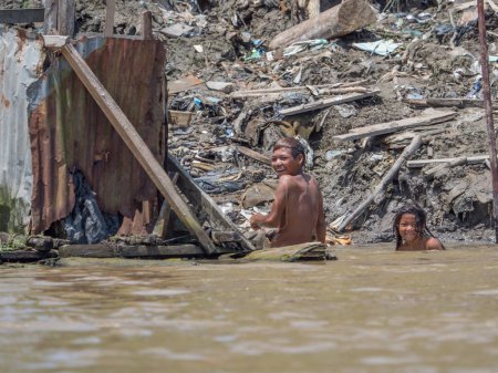 Téléchargez les photos : Iquitos, Pérou - 25 sept. 2018 : Les hommes portent les sacs avec du charbon de bois. Une énorme pollution peut être vue en arrière-plan sur la rive de la rivière Itaya. Basse saison des eaux de l'Amazonie. Quartier Beln d'Iquitos Pérou - en image libre de droit