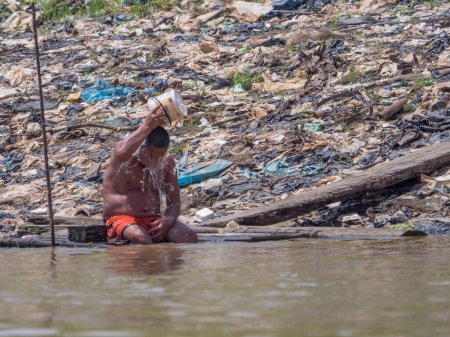 Téléchargez les photos : Iquitos, Pérou - 25 sept. 2018 : L'homme est assis sur les rives de la rivière Itaya et refroidit son corps avec de l'eau. Une énorme pollution peut être vue en arrière-plan. Basse saison des eaux de l'Amazonie. Quartier Beln d'Iquitos Pérou - en image libre de droit