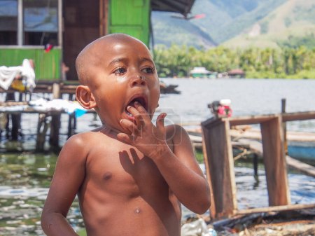 Téléchargez les photos : Sentani, Indonésie - 24 janvier 2015 : Un petit garçon de la tribu Sentani debout devant sa maison, sur la rive du lac Sentatni, au-dessus de l'écorce d'arbre Kombouw peinture art forme - en image libre de droit
