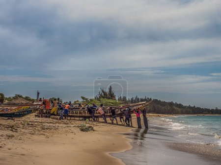 Photo for Nianing, Senegal - January 2, 2019: Fishermen collecting fish from colored wooden fisher boat standing on the beach. Africa - Royalty Free Image