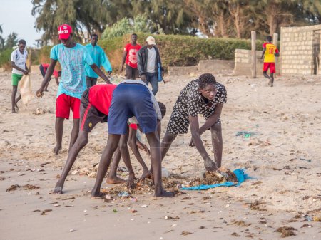 Photo for Senegal, Africa - January 26, 2019:  Young african boys are cleaning the beach from the pollution to play football. Pollution concept. Colorful fisher boats in the background. Senegal. Africa. - Royalty Free Image