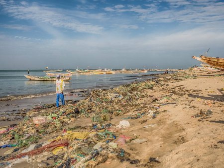 Photo for Senegal, Africa - January 26, 2019:  Black man i walking by the beach with plenty of waste. Pollution concept. Colorful fisher boats in the background. Senegal. Africa. - Royalty Free Image